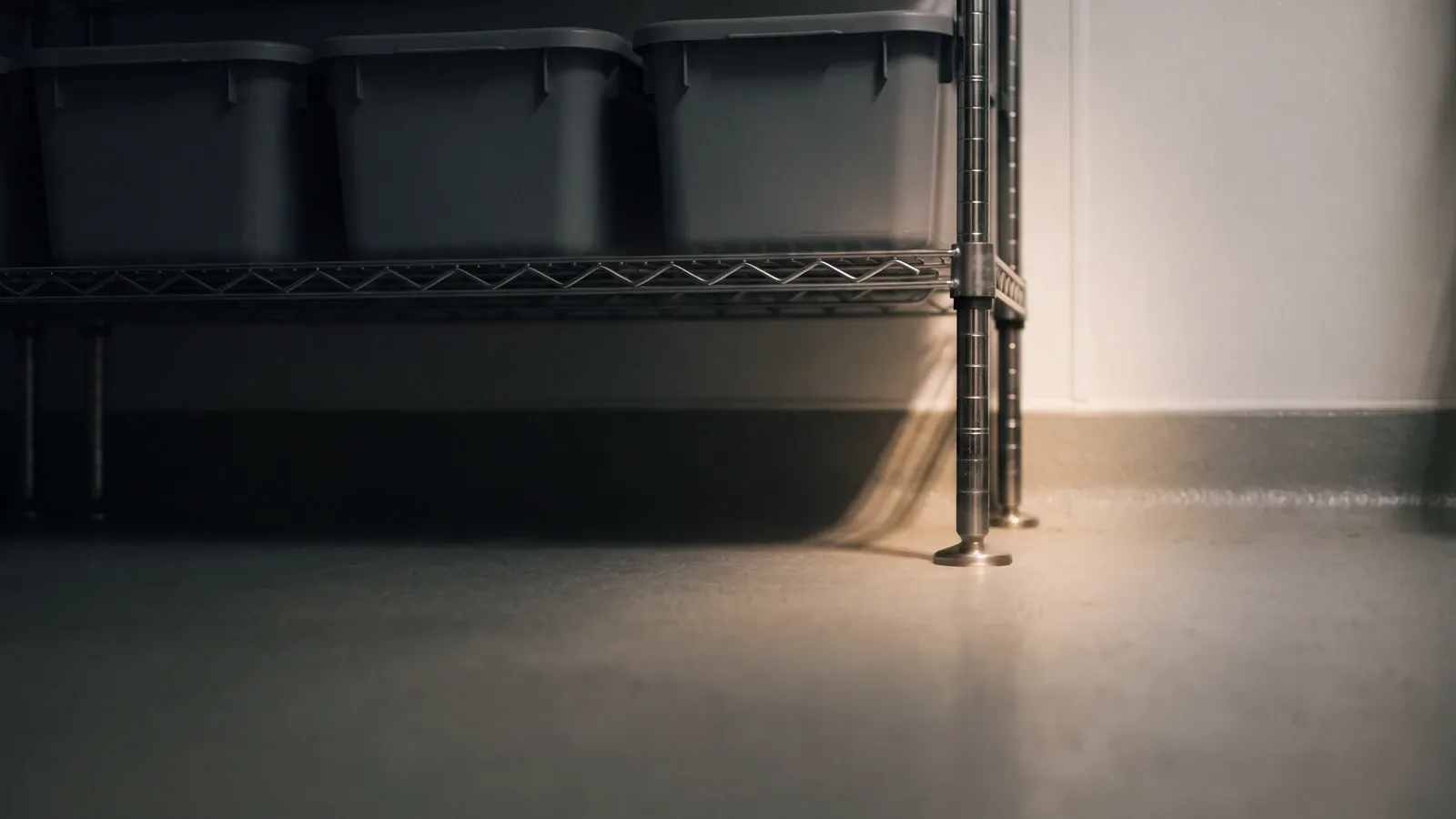 Nahaufnahme in Bodenhöhe in einem dunklen Trockenlager: Edelstahlfuß eines Drahtregals an der Fuge zwischen Boden und Wand, ein schmaler Streiflichtstrahl, darüber neutrale Vorratsbehälter im Schatten.